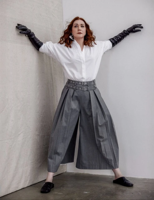 A red-haired woman in an oversized white shirt and gray culottes poses in a photo studio wearing long black leather gloves.