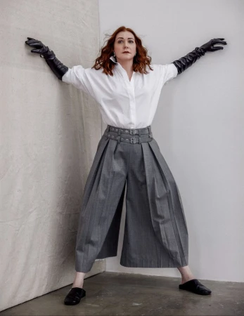 A red-haired woman in an oversized white shirt and gray culottes poses in a photo studio wearing long black leather gloves.