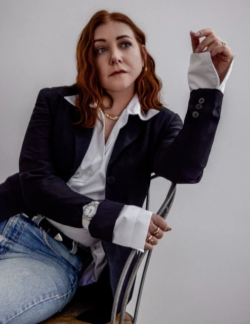 Stylish woman with red hair poses in a navy blazer, jeans, and a white shirt against a neutral background, looking to the side.