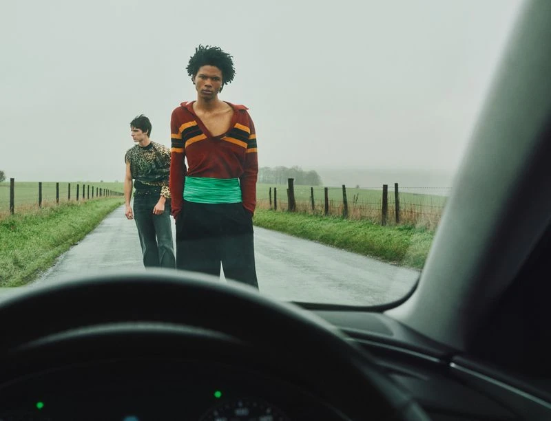 Two fashion models pose on a rural road on a misty day, captured from inside a car, showcasing contemporary clothing styles.