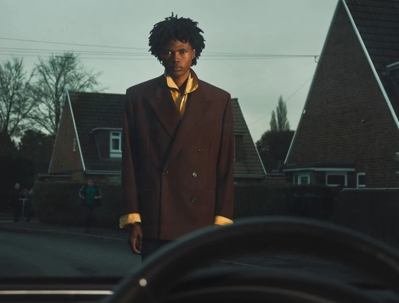 Young man in a brown suit and yellow shirt stands on a street in front of houses, creating a striking and fashionable portrait.