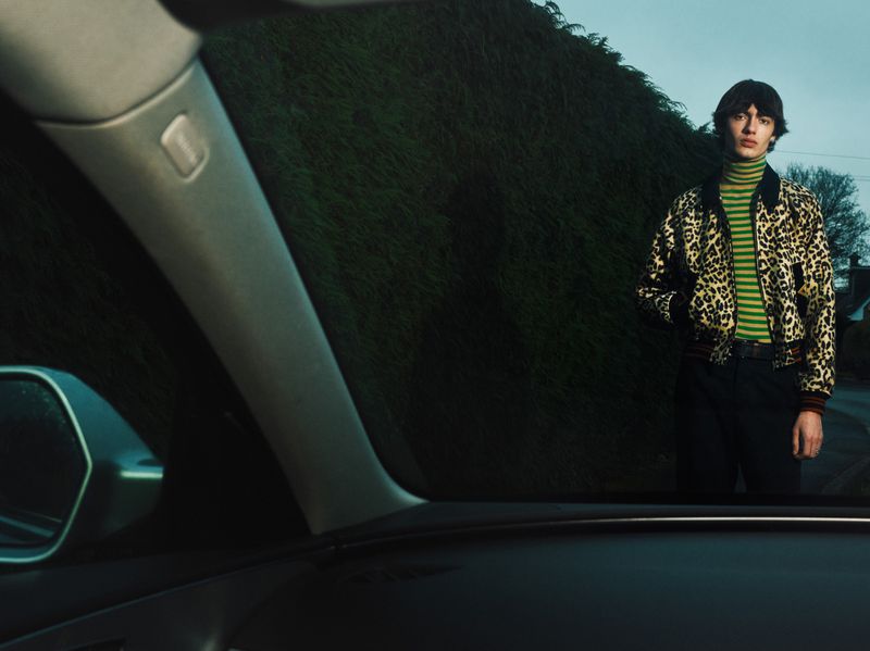 A young adult male poses outdoors wearing a leopard print jacket over a green-striped turtleneck, standing in front of a green hedge.