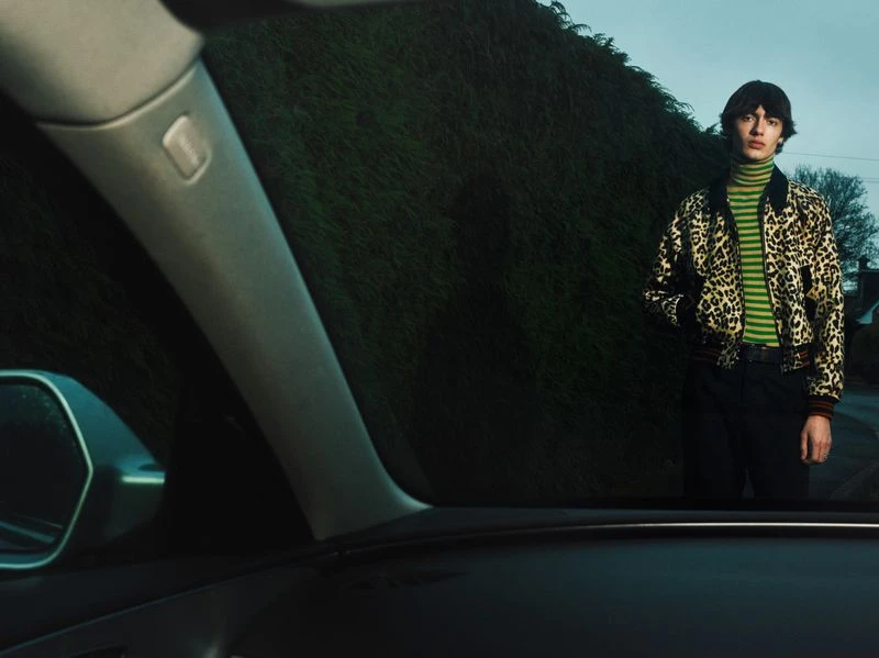 A young adult male poses outdoors wearing a leopard print jacket over a green-striped turtleneck, standing in front of a green hedge.