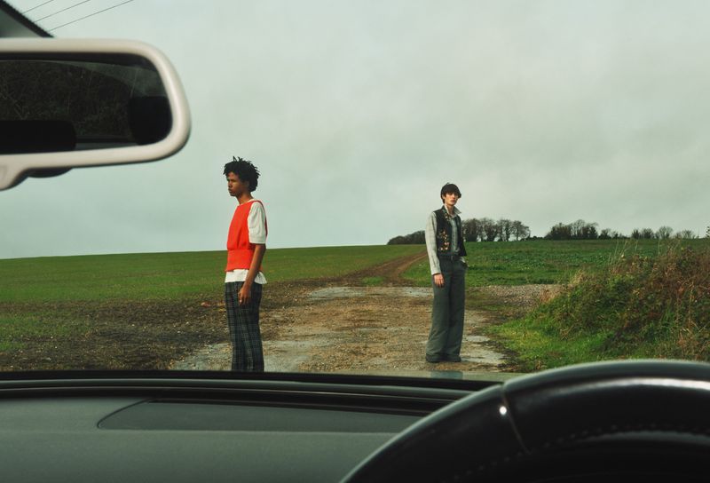 Two adult models stand on a dirt road in a green field under a cloudy sky, viewed from inside a car with visible steering wheel.
