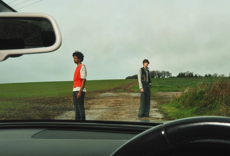 Two adult models stand on a dirt road in a green field under a cloudy sky, viewed from inside a car with visible steering wheel.