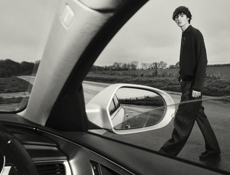 Black-and-white outdoor shot of a male model walking on a rural road, viewed through the windshield of a car on an overcast day.