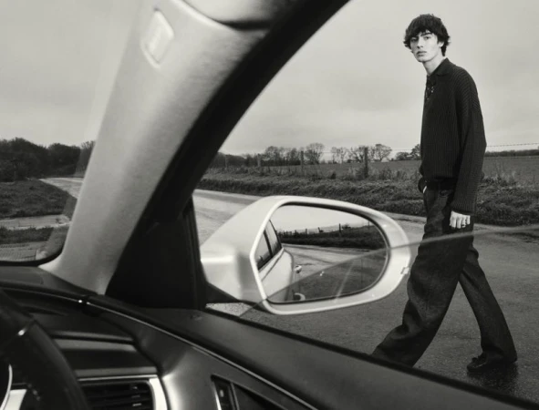 Black-and-white outdoor shot of a male model walking on a rural road, viewed through the windshield of a car on an overcast day.