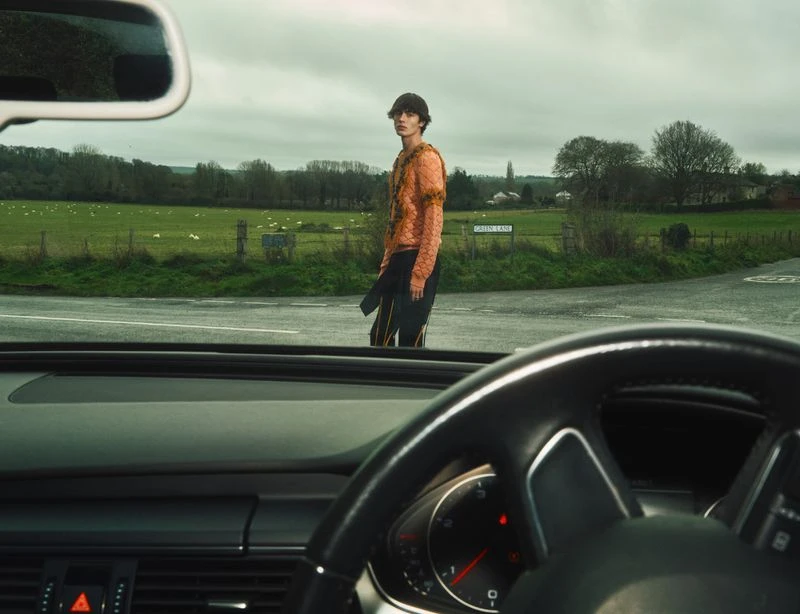 A young adult male in a patterned sweater stands at a rural intersection, viewed through a car windshield with a green field behind him.