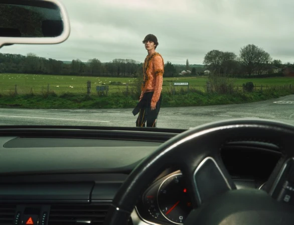 A young adult male in a patterned sweater stands at a rural intersection, viewed through a car windshield with a green field behind him.