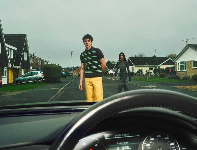 Two people are walking in a residential area on a cloudy day, seen through the windshield of a car. A man and a woman are crossing the street.