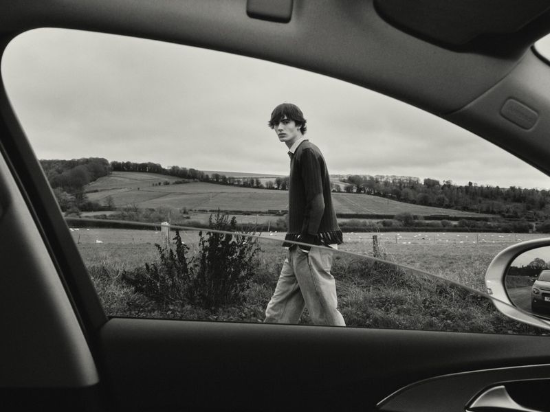 Monochrome view through a car window of a young man walking in an expansive field under an overcast sky, evoking a sense of travel.