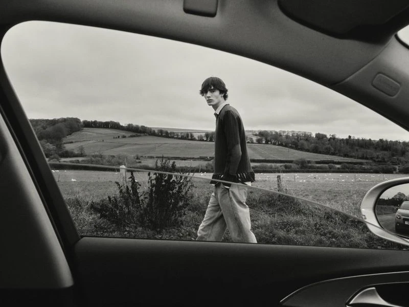 Monochrome view through a car window of a young man walking in an expansive field under an overcast sky, evoking a sense of travel.