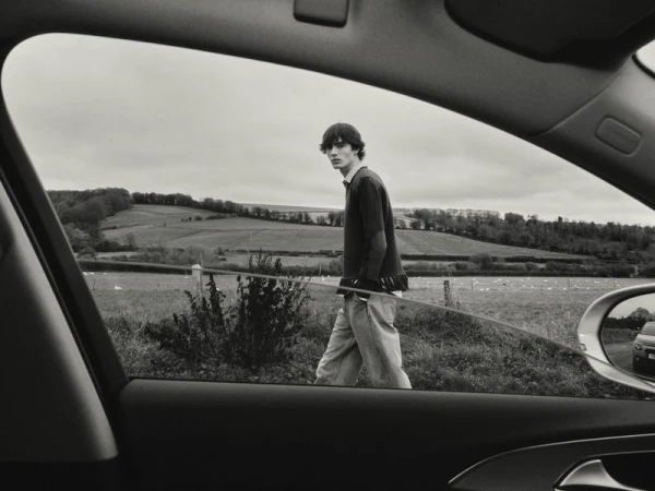 Monochrome view through a car window of a young man walking in an expansive field under an overcast sky, evoking a sense of travel.