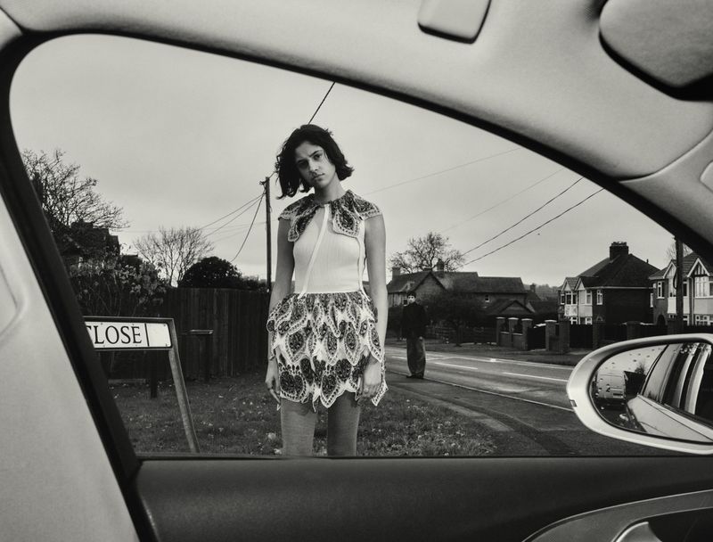 Monochrome shot through car window of woman in patterned dress on street next to 'Close' sign, with house and sky background.