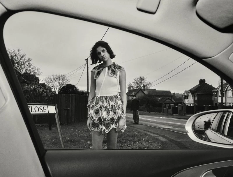 Monochrome shot through car window of woman in patterned dress on street next to 'Close' sign, with house and sky background.
