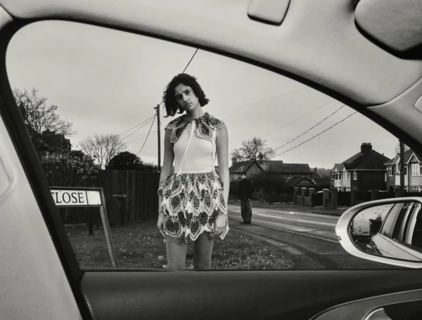 Monochrome shot through car window of woman in patterned dress on street next to 'Close' sign, with house and sky background.