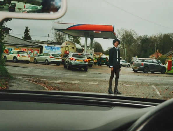 Young adult in a suit stands at a British petrol station. Cars are queued behind him. The shot is taken from inside a car.