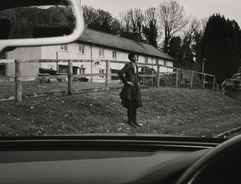 Monochrome photograph from inside a car, showing a person in patterned coat standing outside a rural home with rolling grassy hills.