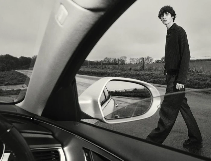 Monochrome fashion editorial image of a young male model walking outside, viewed through the open window of a car.