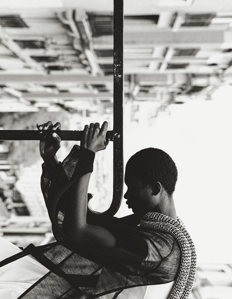 Monochrome portrait of an androgynous person grasping metal bars, with an upside-down building visible in the background, creating a surreal effect.