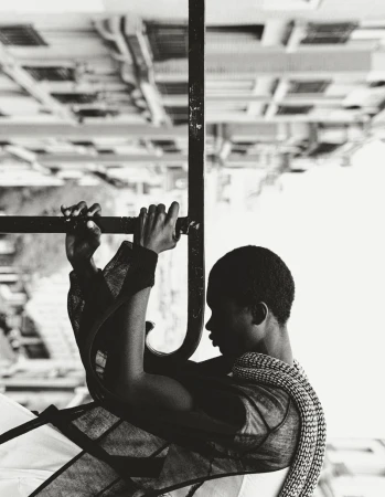 Monochrome portrait of an androgynous person grasping metal bars, with an upside-down building visible in the background, creating a surreal effect.