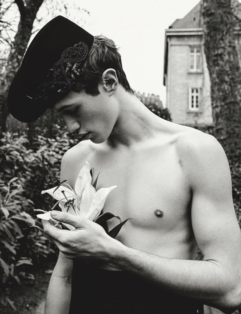 Monochrome portrait of a young man with bare chest, wearing a decorated hat, holding a lily, outdoors with building in the background.