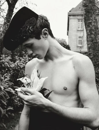 Monochrome portrait of a young man with bare chest, wearing a decorated hat, holding a lily, outdoors with building in the background.