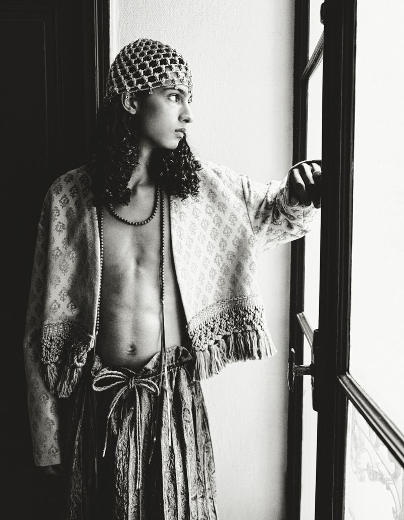 Model with long dark hair wearing a patterned jacket and beaded cap standing by a window for HERO Magazine.