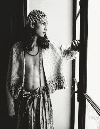Model with long dark hair wearing a patterned jacket and beaded cap standing by a window for HERO Magazine.