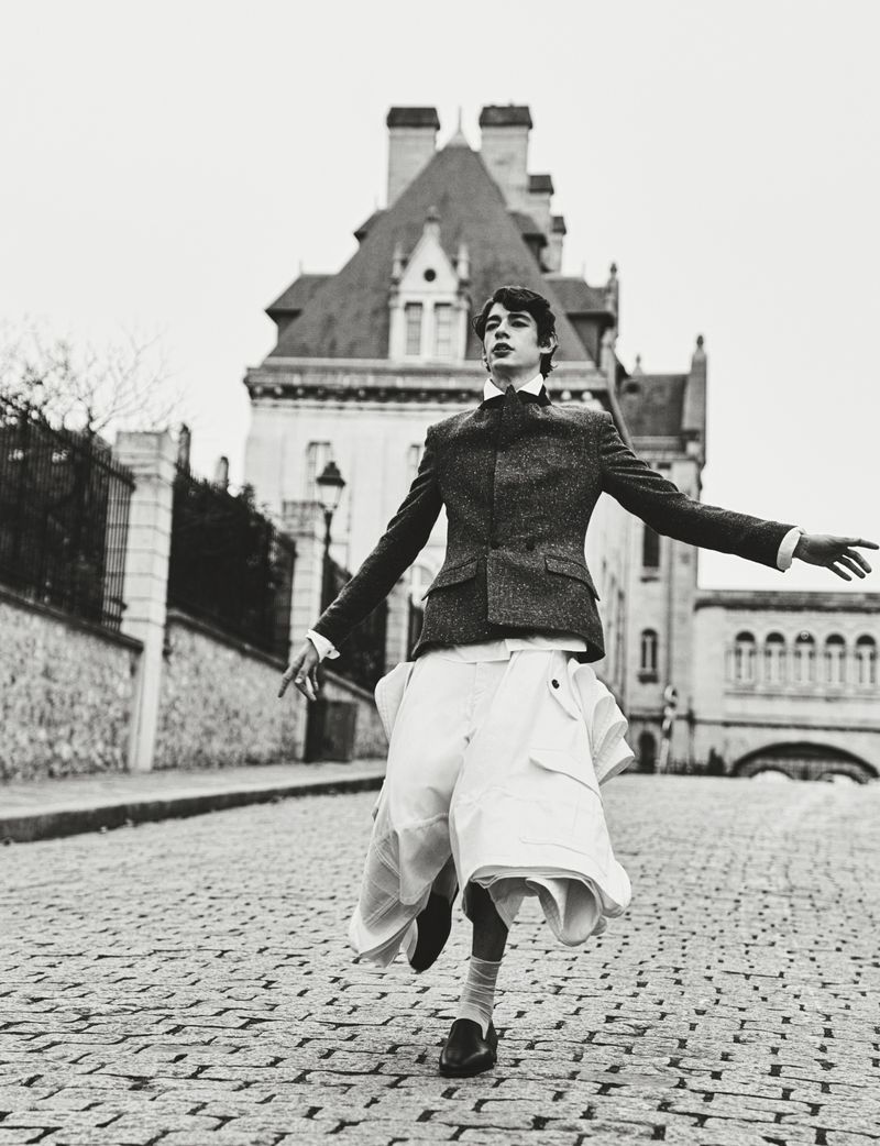 Monochrome outdoor fashion shot of a young adult male model in a blazer, skirt, and dress shoes, on a cobblestone street with a building.