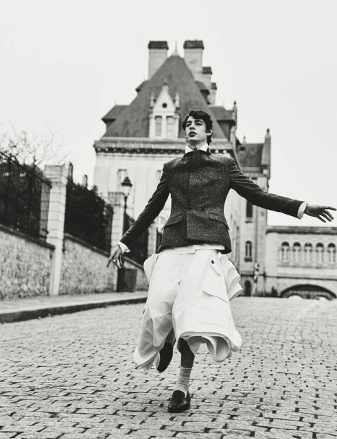 Monochrome outdoor fashion shot of a young adult male model in a blazer, skirt, and dress shoes, on a cobblestone street with a building.