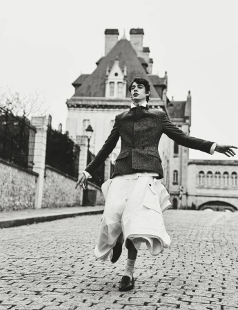 Monochrome outdoor fashion shot of a young adult male model in a blazer, skirt, and dress shoes, on a cobblestone street with a building.