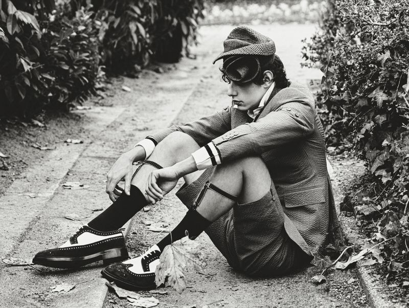 Young adult male in tweed suit sits on pathway wearing spectator shoes in outdoor setting with leafy background. Black and white, editorial.
