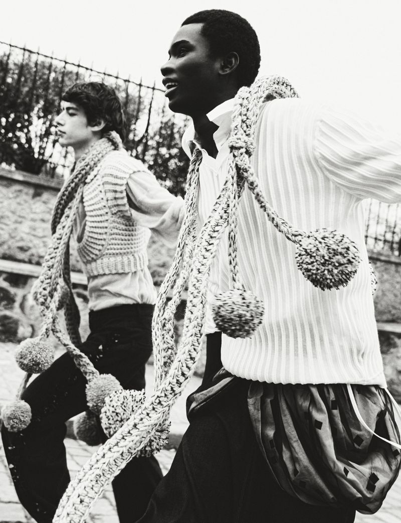 Two young people in black and white wearing knitted vests and rope scarves outside by a stone wall under a cloudy sky.