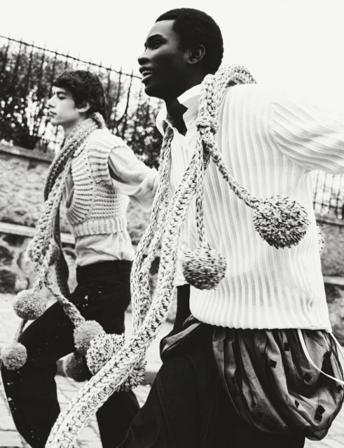 Two young people in black and white wearing knitted vests and rope scarves outside by a stone wall under a cloudy sky.
