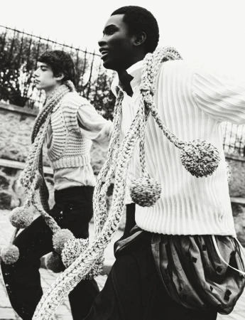 Two young people in black and white wearing knitted vests and rope scarves outside by a stone wall under a cloudy sky.