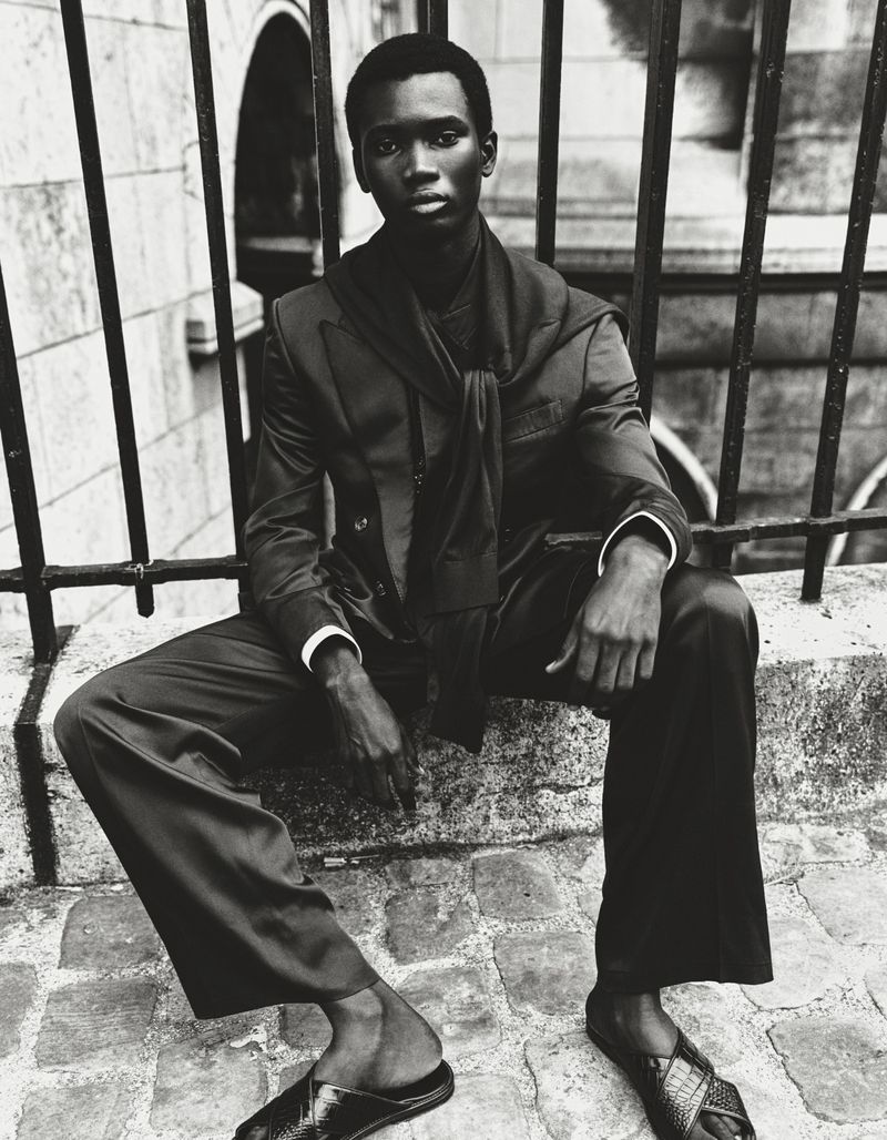 Monochrome portrait of a Black male model sitting on a ledge in a suit and slides, leaning against metal railings, outdoor fashion shot.