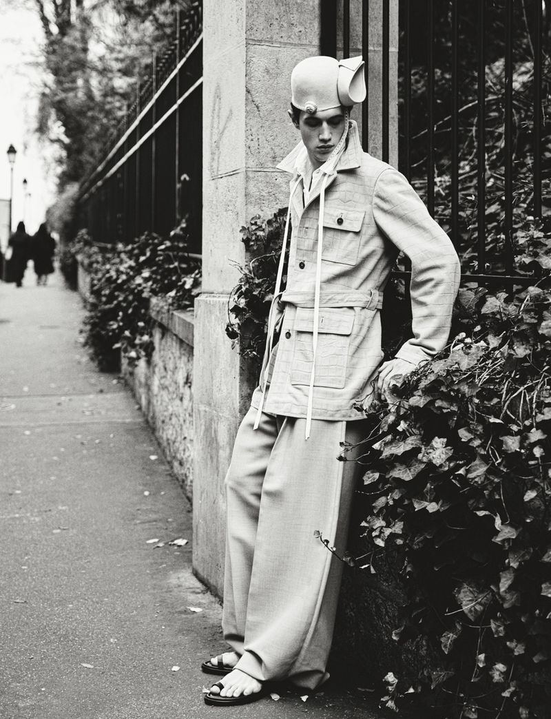 Monochrome fashion shot of a male model in an urban setting wearing wide-leg pants, a light jacket, sandals, and an unusual hat.