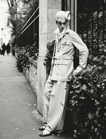 Monochrome fashion shot of a male model in an urban setting wearing wide-leg pants, a light jacket, sandals, and an unusual hat.