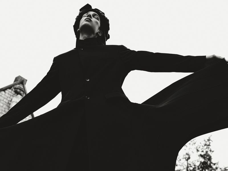 Monochrome fashion shot of a slender person in an open, buttoned coat with arms outstretched against a bright sky and background building.