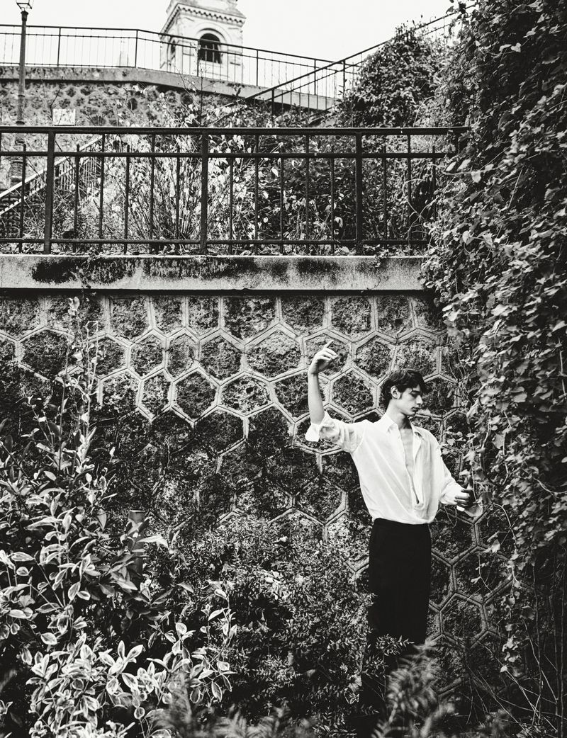 Monochrome portrait of a stylish man posing outdoors near a stone wall and lush plants, captured in a fine art photograph.