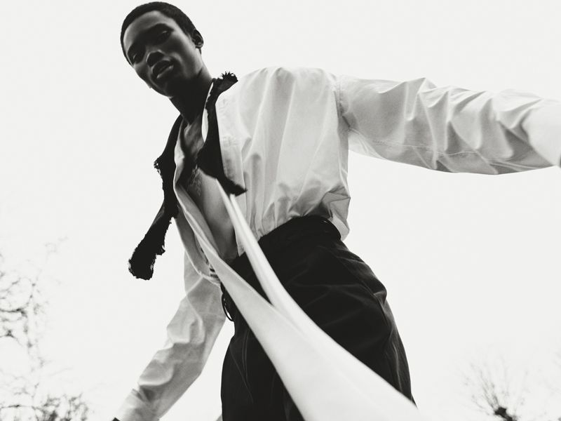 Low-angle shot of an African male model in a white shirt and dark pants against a bright sky. Fashion editorial with dramatic lighting.