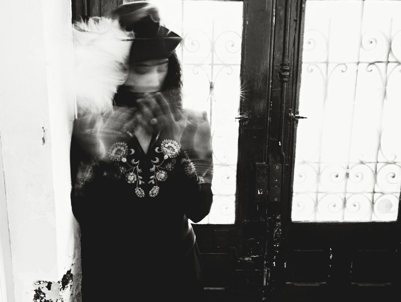 Monochromatic portrait of a blurry person wearing a hat and an embroidered shirt, standing near a dark wooden door with ornate glass.