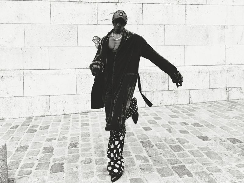 A Black man wearing a robe and patterned boots runs on a stone-paved street with a neutral stone building behind him in black and white.