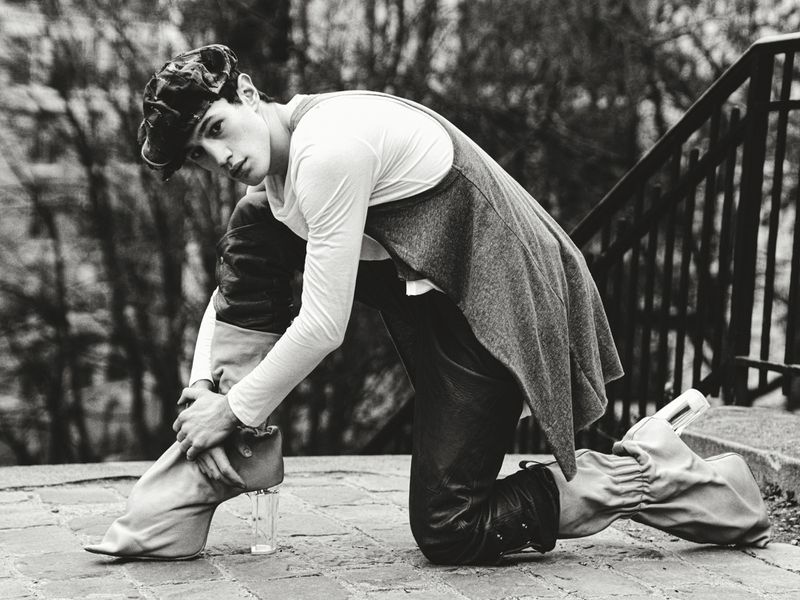 Monochrome fashion shot of a model kneeling on cobblestone in leather pants, a white long sleeve shirt, gray vest, cap, and glass-heeled boots.