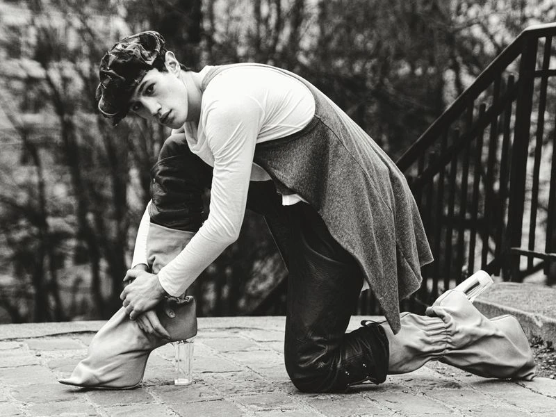 Monochrome fashion shot of a model kneeling on cobblestone in leather pants, a white long sleeve shirt, gray vest, cap, and glass-heeled boots.