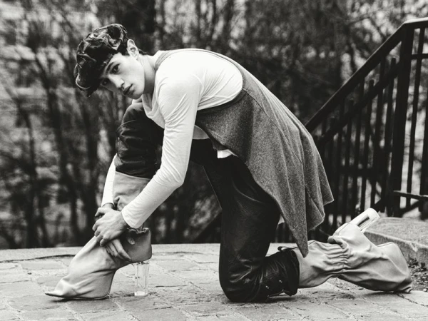Monochrome fashion shot of a model kneeling on cobblestone in leather pants, a white long sleeve shirt, gray vest, cap, and glass-heeled boots.