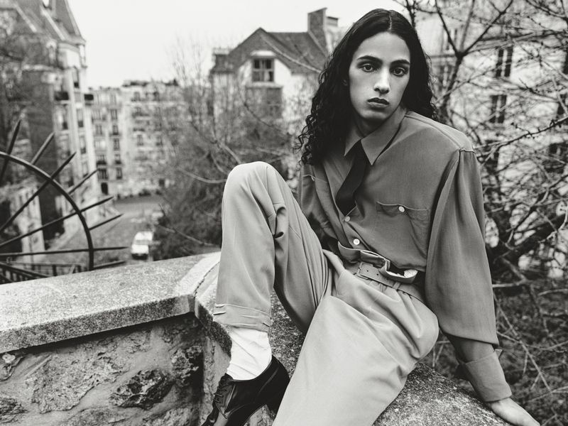 Young person with long dark hair, wearing a button-down shirt and light-colored trousers, posing on a stone wall in an urban setting.