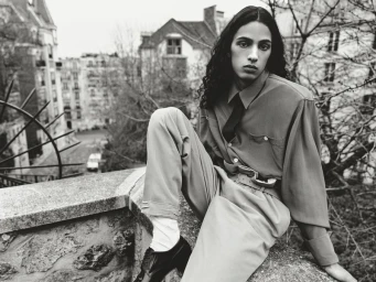 Monochrome fashion editorial portrait of a model sitting on a stone wall with a city street background for HERO Magazine