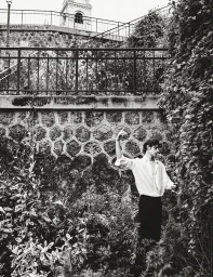 Black and white editorial image of a male model in a white shirt standing near stone walls and lush foliage.